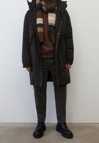 Zwarte parka met capuchon, bordeaux sweatshirt, donkere broek en zwarte chunky laarzen. Gestreepte bruine en groene gebreide sjaal als accent.
