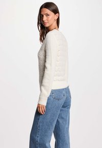 Weißer, flauschiger Pullover mit strukturiertem Zickzackmuster auf dem Rücken, kombiniert mit hellblauen Jeans. Lockere Passform, lange Ärmel und gerippte Bündchen.