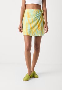 Falda envolvente en azul claro con rayas amarillas, con detalle de lazo en la cintura. Textil suave, de longitud por encima de la rodilla. Combinada con mocasines verdes.