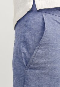 Pantalones de lino azul con un acabado texturizado, que presentan un bolsillo lateral y un corte entallado, parcialmente visibles debajo de una prenda blanca.