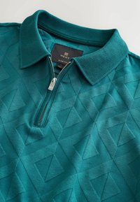 Slim fit polokošile v teal barvě, s oboustranným límcem, strukturovaným geometrickým vzorem a měkkou tkaninou. Nálepka značky je viditelná uvnitř.