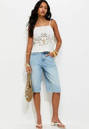 Femme portant un haut sans manches blanc brodé, un short bermuda en denim bleu clair, des sandales blanches, et portant un sac tissé décoré de coquillages.