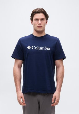 Jeune homme aux cheveux bruns courts, portant un t-shirt Columbia bleu marine et un pantalon gris, debout devant un fond clair uni.