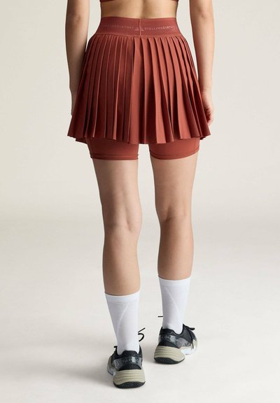 Rückansicht einer Person, die einen rostfarbenen, plissierten Tennisshortrock mit integrierten Shorts, weiße Wadenstrümpfe und schwarze Sportschuhe trägt.