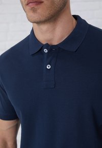 Navy blue polo shirt made of textured fabric with a collar, two buttons, and a subtle pattern. Close-up view highlighting details.