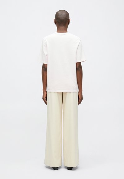 Osoba s oholenou hlavou, oblečená v růžovém tričku s krátkým rukávem, širokých krémových kalhotách a černých botách, stojící zády na bílém pozadí.