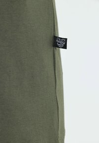Camisa de algodón verde olivo con costura lateral, que presenta una etiqueta tejida negra con texto blanco. Textura suave, diseño casual.