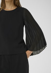 Top nero con maniche lunghe, trasparenti e plissettate. Collo rotondo, corpo in tessuto liscio e vestibilità morbida. Design semplice che mette in risalto i dettagli delle maniche.