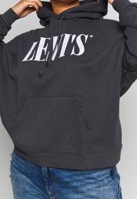 Personne portant un sweat à capuche Levi's gris foncé avec un logo blanc et un jean en denim bleu, ajustant la capuche avec les deux mains levées.