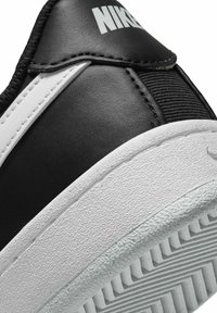 Baskets en cuir synthétique noir avec une semelle en caoutchouc blanche texturée, arborant un logo Nike bien visible et un détail d'une bande blanche contrastante.