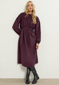 Abito lungo borgogna a maniche lunghe con corpetto arricciato, vita cinturata e silhouette fluida, indossato con stivali neri fino al ginocchio. Tessuto liscio.