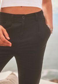 Pantaloni neri con un design aderente, dotati di chiusura con bottone, pieghe e tasche laterali. Tessuto dalla trama liscia.