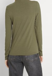 Olivgrünes, geripptes Langarmshirt mit hohem Kragen, das eine glatte Textur und eine taillierte Form aufweist, kombiniert mit dunkelgrauen Jeans.