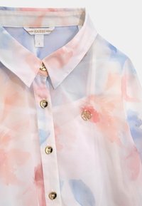 Chemisier transparent aux teintes pastel de rose et de bleu, avec des boutons dorés et une épingle décorative portant un logo au niveau de la poitrine.