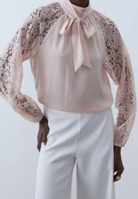 Blusa realizzata in morbido tessuto rosa, con maniche in pizzo, collo alto e un dettaglio a fiocco al collo, abbinata a pantaloni bianchi.