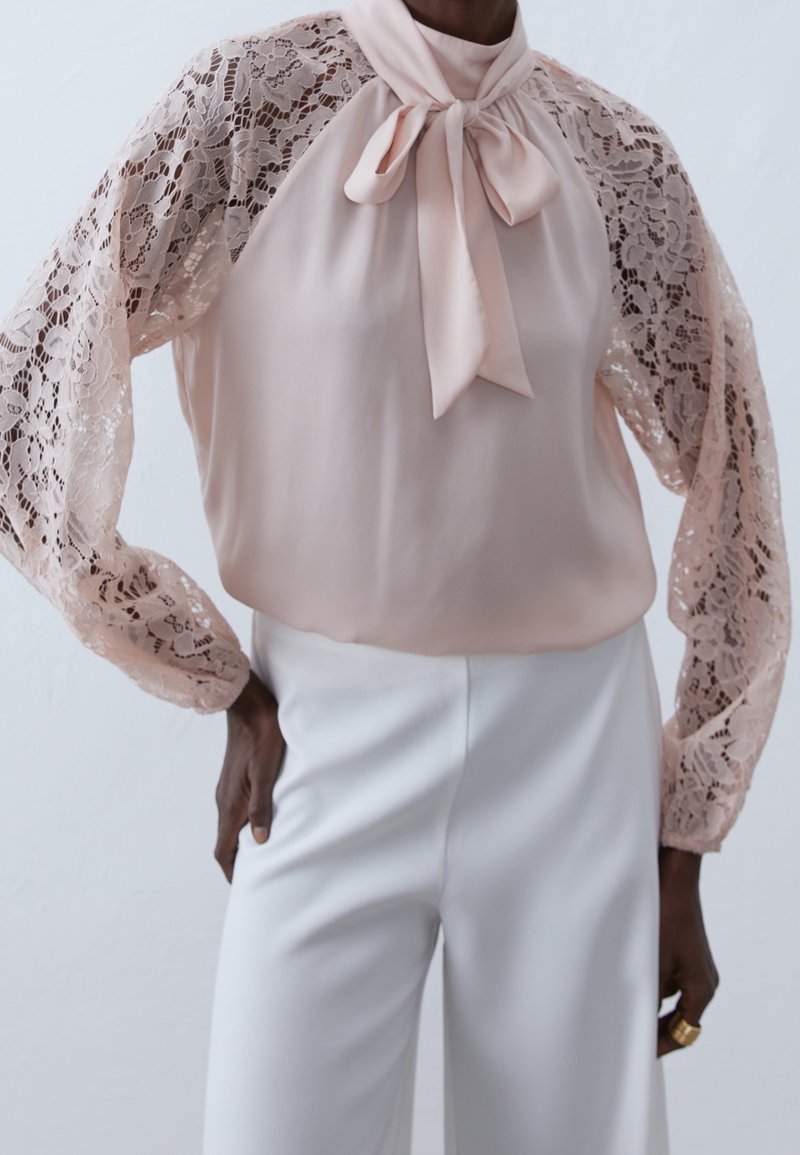 Blusa realizzata in morbido tessuto rosa, con maniche in pizzo, collo alto e un dettaglio a fiocco al collo, abbinata a pantaloni bianchi.