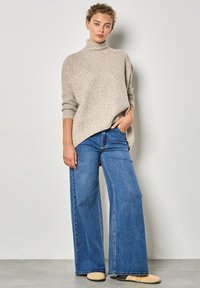 Maglione a dolcevita beige macchiato con una vestibilità comoda, abbinato a jeans blu a gamba larga. Pantofole marrone chiaro visibili. Design semplice e informale.