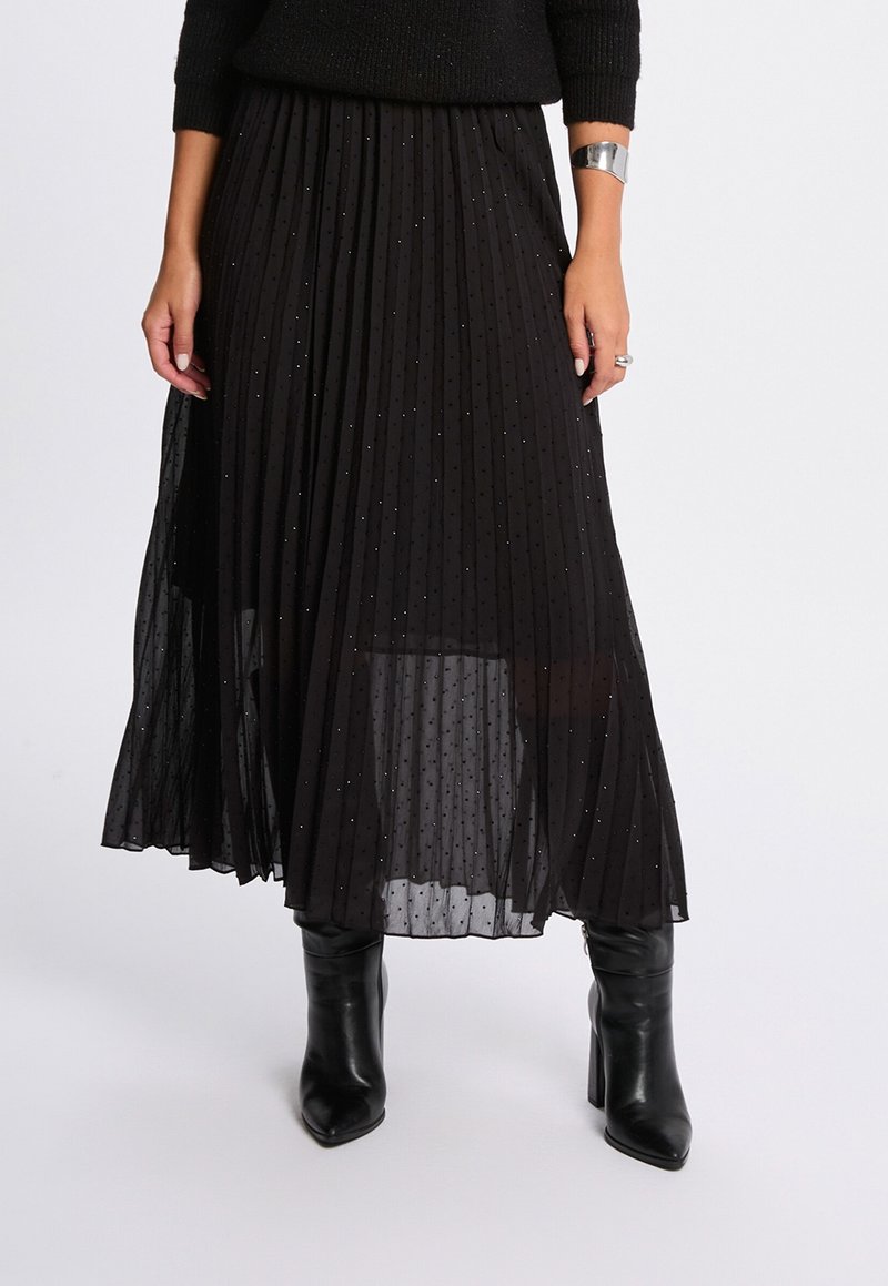Femme portant une jupe midi plissée noire avec des paillettes subtiles, des bottines noires à bout pointu et talons, et un bracelet manchette argenté, debout devant un fond blanc.