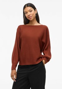 Rostbrun ribbadsweater med bred rundhalsning och långa ballongsärmar. Kombinerad med svarta byxor för kontrast. Texturerad yta.