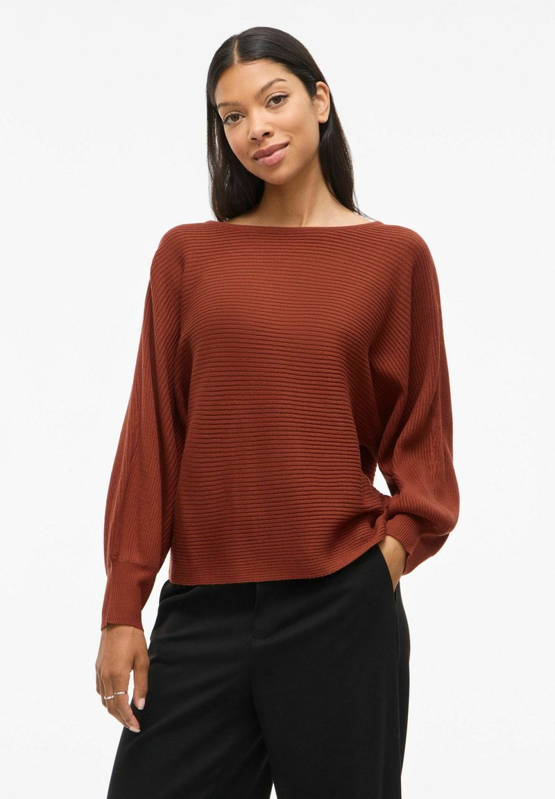 Rostbrun ribbadsweater med bred rundhalsning och långa ballongsärmar. Kombinerad med svarta byxor för kontrast. Texturerad yta.