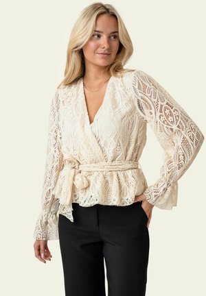 Kobieta ubrana w kremową koronkową bluzkę z długimi rękawami i wiązaniem w talii, w połączeniu z czarnymi spodniami, na neutralnym tle.