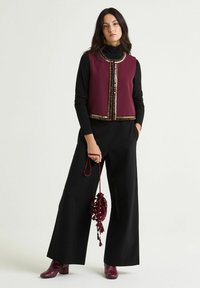 Gilet senza maniche color bordeaux con perline decorative, indossato sopra un dolcevita nero. Pantaloni neri a gamba larga e stivaletti bordeaux con texture completano il look.