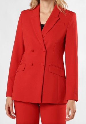 Vrouw draagt een op maat gemaakte rode double-breasted blazer en bijpassende rode broek, met een deels zichtbare zwarte top eronder.