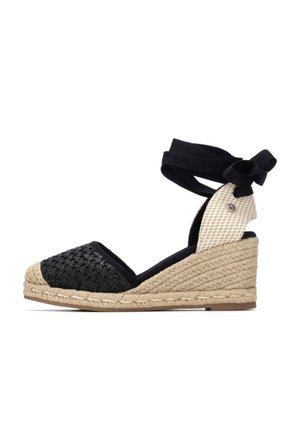 Alpargata cuña de tela negra tejida con suela de yute trenzado y atadura en el tobillo. Cuenta con un panel de tacón con patrón blanco y negro y un detalle decorativo.