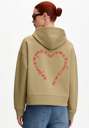 Roodharige vrouw met knot draagt een beige hoodie met rode hartvormige tekst op de rug en blauwe jeans, zijwaarts kijkend tegen een witte achtergrond.