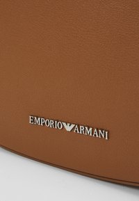 Brun tekstureret lædertaske med sølv "Emporio Armani" logo. Glat overflade og rund form, med et minimalistisk design.