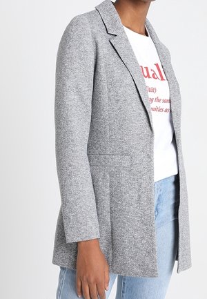 Vrouw draagt een lichtgrijze getextureerde blazer over een wit grafisch T-shirt en lichtblauwe spijkerbroek, getoond van schouder tot halverwege de dij.