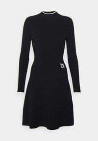 Robe en tricot noir à côtes avec des manches longues, un col montant, un corps ajusté, une jupe évasée et un détail de logo blanc sur le côté gauche.
