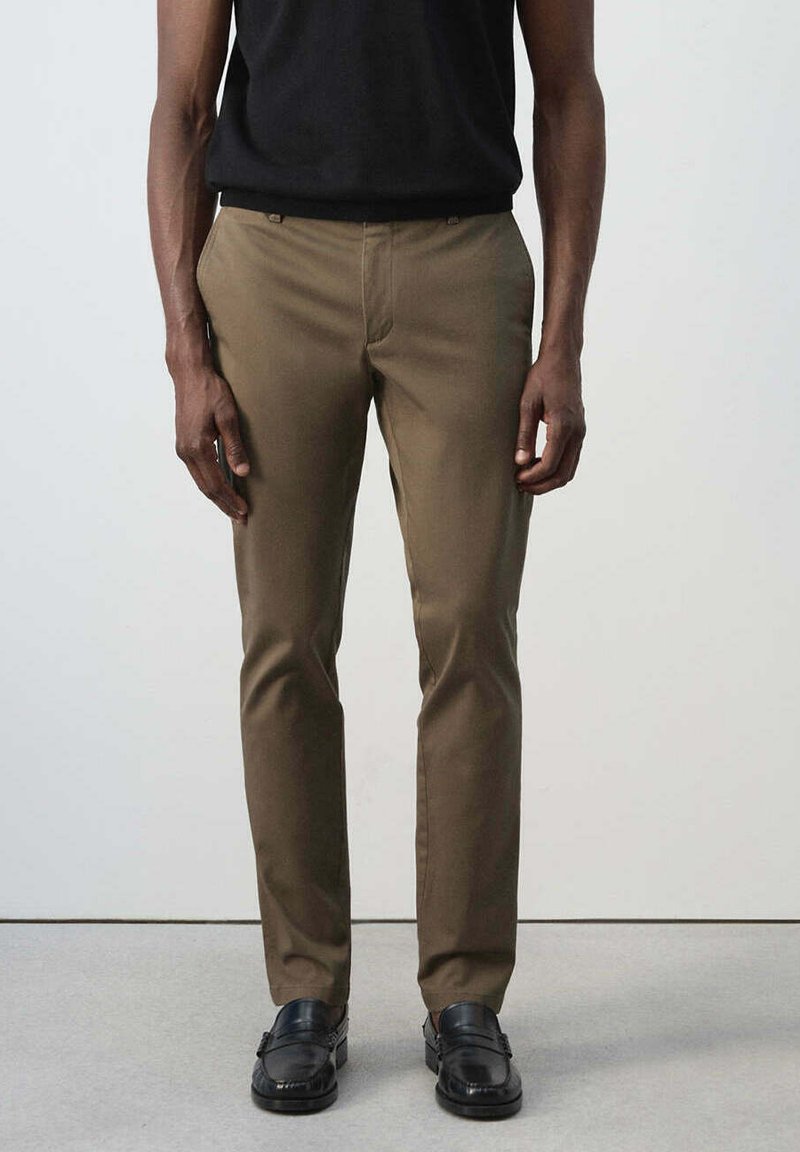 Homme portant un pantalon marron slim et des mocassins en cuir noir, debout devant un fond uni.