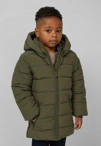 Mali dječak kratke kovrčave kose nosi maslinasto zeleni puffer kaput s kapuljačom i rukama u džepovima na jednostavnoj pozadini.
