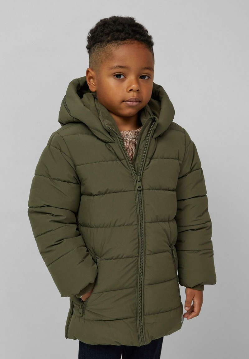 Mali dječak kratke kovrčave kose nosi maslinasto zeleni puffer kaput s kapuljačom i rukama u džepovima na jednostavnoj pozadini.