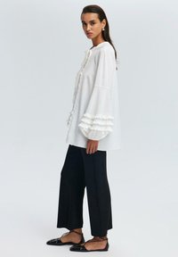 Hvid, rynket bluse med lange, udvidede ærmer, parret med sorte bukser med brede ben og sorte flade sko med stropper. Simpelt, elegant design.