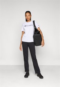 Weißes T-Shirt mit blauem "Champion"-Logo, schwarze Hose und schwarze Schuhe; hält eine schwarze Tasche mit Schultergurt; elegantes Design, lässiger Stil.