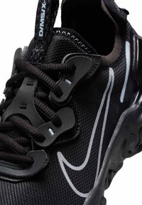 Zapatilla deportiva negra con parte superior de malla, cordones elásticos y suela texturizada. Detalles reflectantes y un diseño distintivo de la marca en blanco.