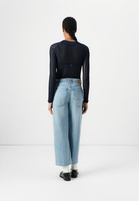 Maglione lungo a maniche nere in maglia con un motivo testurizzato, abbinato a pantaloni larghi in denim azzurro chiaro. Scarpe nere e calzini bianchi visibili.