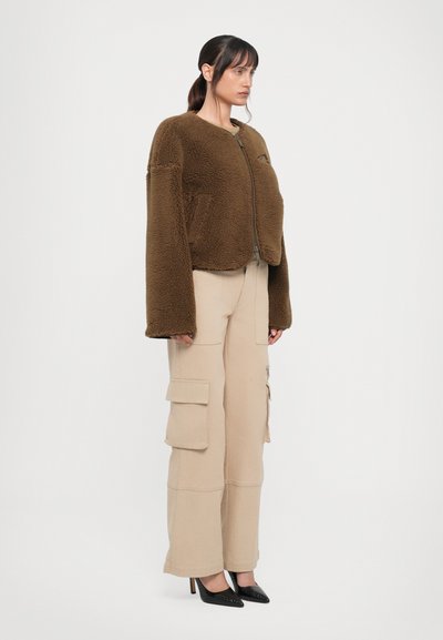 Braune Fleecejacke mit Rundhalsausschnitt, langen Ärmeln und Reißverschluss. Kombiniert mit beigen Cargo-Hosen und schwarzen High Heels.