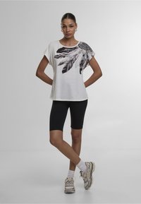 Camiseta blanca de manga corta con estampado de plumas negras en el hombro izquierdo, combinada con pantalones cortos de ciclismo negros y zapatillas deportivas grises.