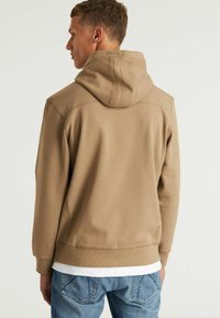 Felpa beige con cappuccio e finitura testurizzata, dotata di tasca a marsupio e polsini a coste, abbinata a jeans azzurri chiari.