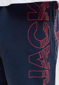 Donkerblauwe shorts met dikke rode, gestreepte tekst "NAVY" die verticaal langs één zijde is gedrukt, gedeeltelijk bedekt door een wit kledingstuk.