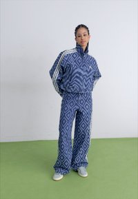 Blauw geoordeeld trainingspak met een ritsjack en wijde broekspijpen. Heeft witte strepen langs de mouwen en benen, aangevuld met lichte sneakers.