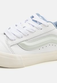 White low-top sneaker with textured sole, white laces, light gray side stripe, and light blue padded collar.