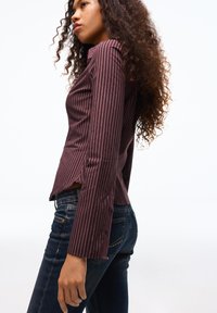 Bordowa bluzka z długim rękawem w białe pionowe paski, o dopasowanym kroju, z bocznymi rozcięciami i guzikami na mankietach, zestawiona z ciemnymi jeansami.