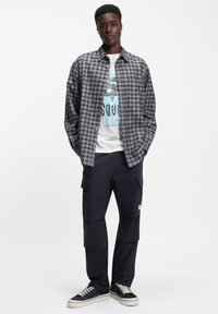 Harmaa ruudullinen napillinen paita valkoisen graafisen t-paidan päällä, yhdistetty mustiin cargo-housuihin ja mustiin lenkkareihin, joissa on kermaiset nauhat.