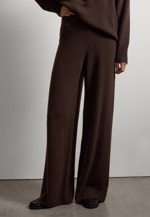 Person iført løse, mørkebrune strikkede vidde bukser med matchende sweater og sorte slip-on sko, stående på en grå gulv.