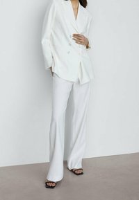 Blazer blanc à double boutonnage associé à un pantalon blanc évasé et des sandales à talons noires ; tissu lisse, design ajusté, détails minimalistes.