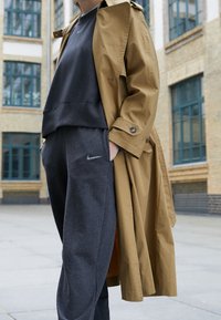 Brun trenchcoat över en grå sweatshirt och mörkgrå sweatpants. Noterbara funktioner inkluderar knappar och en synlig logotyp.